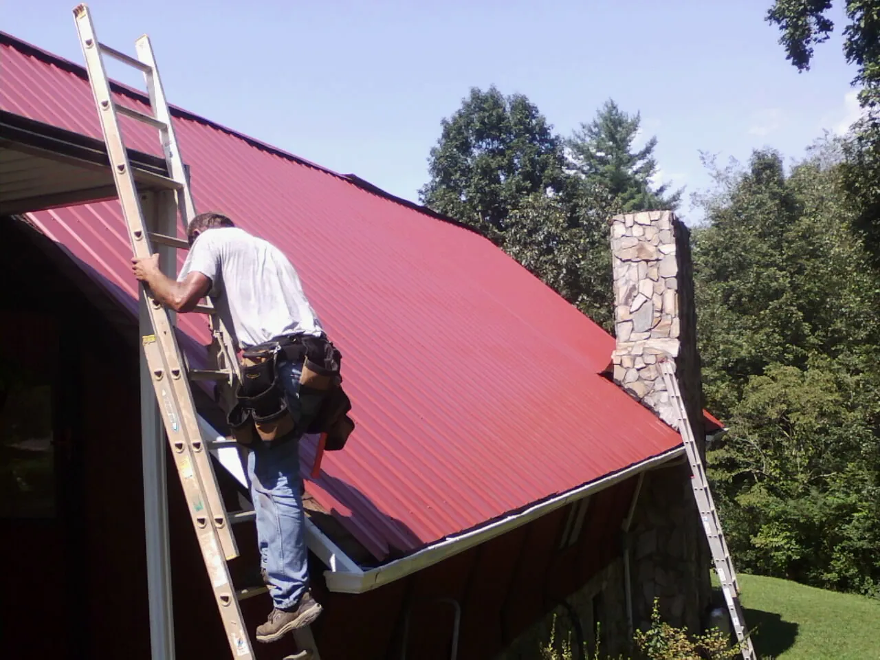 Roofer on a ladder beside a red metal roof and stone chimney — Soffit & Fascia Repair by Roofing Brothers in Sterling